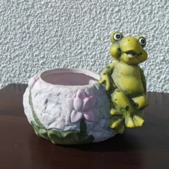 Γλαστρούλα  που απεικονίζει έναν χαμογελαστό βάτραχο να κάθεται αναπαυτικά δίπλα σε ένα ανθισμένο νούφαρο.