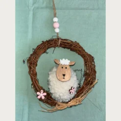αρνάκι με μαλακό σώμα και ξύλινο κεφάλι. Ένα φυσικό στεφάνι από κλαδιά αγκαλιάζει τη σύνθεση. Μικρά ροζ λουλούδια και ένας φιόγκος από ψάθα στολίζουν το κάτω μέρος