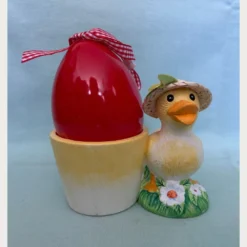 κίτρινο παπάκι που φοράει ψάθινο καπελάκι και στέκεται σε μια ανθισμένη βάση. Η θήκη περιλαμβάνει ένα μεγάλο κόκκινο κερί σε σχήμα αυγού με καρό κορδέλα.